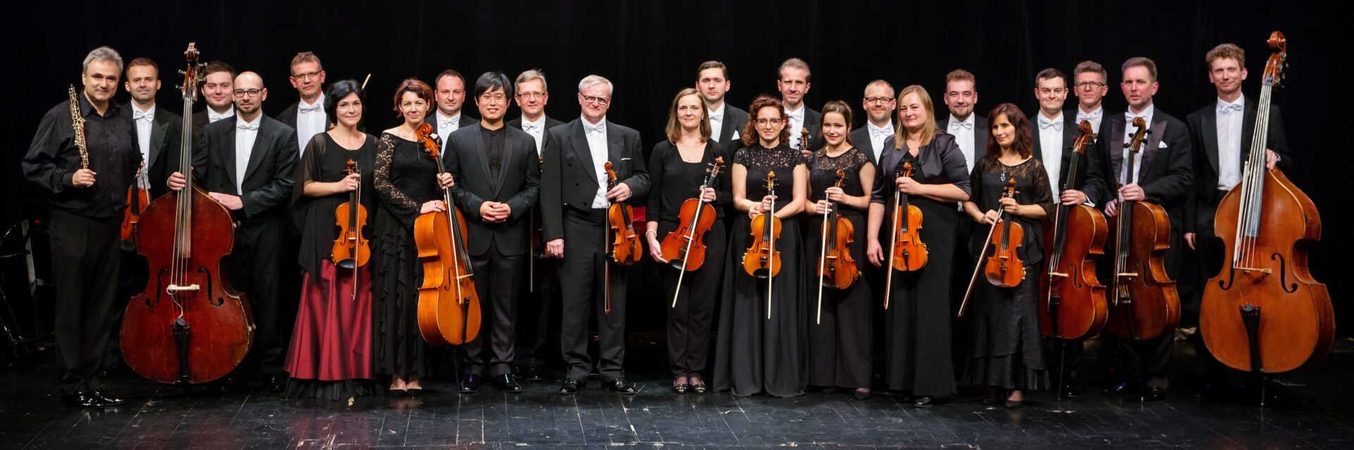 zdjęcie prezentujące muzyków Gliwickiej Orkiestry Kameralnej stojących na scenie. Wszyscy elegancko ubrani z instrumentami w rękach.